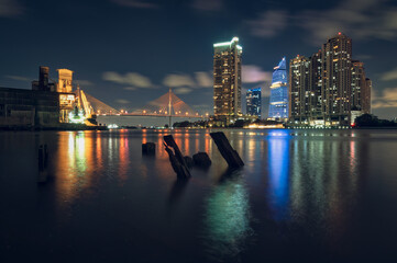Obraz premium Scenic night cityscape of riverbank with old wooden stumps in water and illuminated city buildings background.