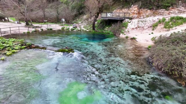 Aerial tracking shot flying forward over the crystal-clear turquoise waters of the Blue Eye spring toward the wooden observation deck and viewing platform in Albania