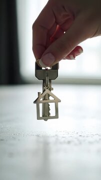 Extreme close-up video of a modern, gender-ambiguous hand with clean nails placing a sleek silver key with a house-shaped keychain onto light-grey concrete, dolly-in. Concept of new beginnings and