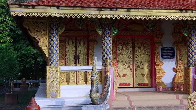 Low-altitude drone shot tracks sideways across the ornate facade of a traditional Buddhist temple pavilion, showcasing intricately carved golden doors, decorated pillars, and protective Naga serpent s