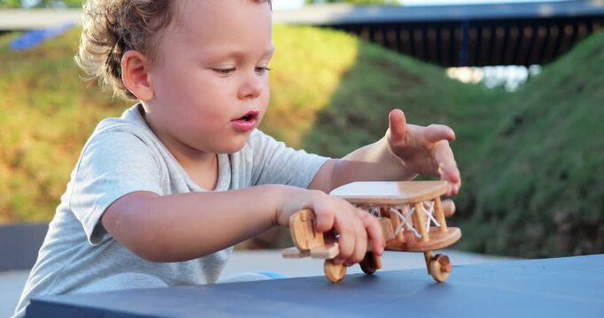Toddler plays with wooden toy biplane, holding it with one hand while using other to spin propeller of tiny toy. His open-mouthed fascination and delight are evident as he watches propeller turn.