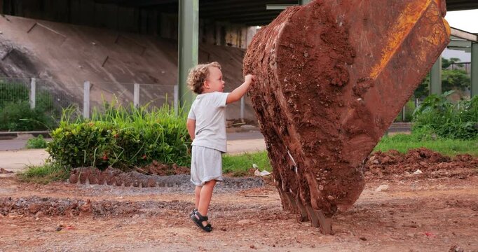 Curious toddler, holding small stone, scrapes dried dirt from large bucket of parked excavator. Engaging in tactile exploration, baby boy fearlessly examines huge piece of machinery