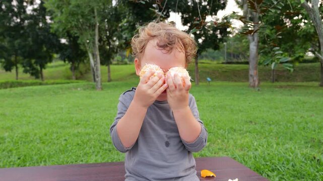 Funny toddler sits on park bench playing peekaboo with peeled mandarins over his eyes, giggling and having innocent fun. Little joker hides his face with fruits, then joyfully reveals himself