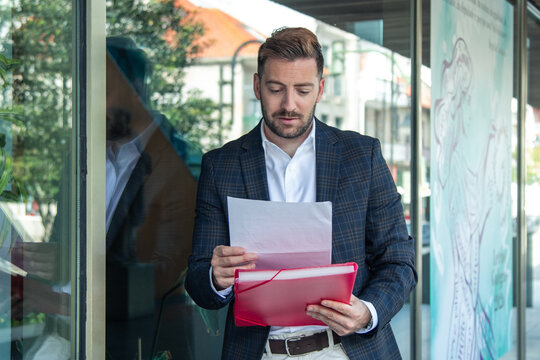 Young professional businessman in a sleek suit reviewing reports and documents on a clipboard while standing outside a contemporary glass corporate building