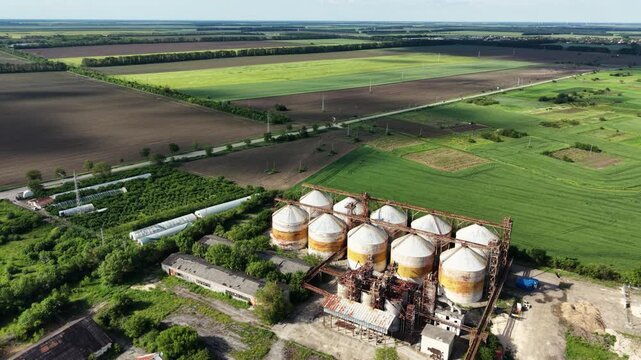 Aerial view of industrial grain silos surrounded by vast agricultural fields and green meadows under a clear sky in Balchik, Dobrich Province, Bulgaria.