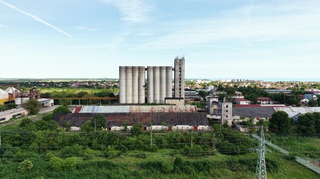 Aerial view of large concrete grain silos and industrial buildings surrounded by lush green fields and power lines under a clear blue sky in Balchik, Dobrich Province, Bulgaria.