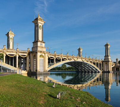 Ornate Seri Gemilang Bridge in Putrajaya, Malaysia, featuring grand towers and golden decorations, beautifully reflected in the calm water under a clear blue sky