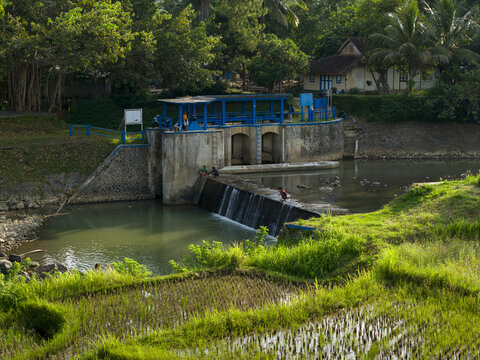 Aerial view of Tangsi Water Gate with a small waterfall, people by the river, and lush green rice fields in Kecamatan Slawi, Jawa Tengah, Indonesia.