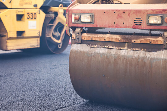 Workers are engaged in asphalt paving on a city street using heavy machinery. The scene showcases the process of road construction in an urban area under clear skies.