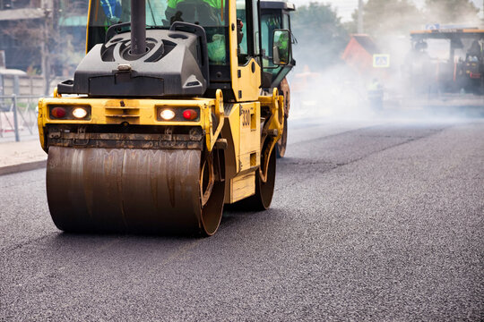 Heavy machinery is engaged in asphalt paving work on a city street. Workers are present as the roller compacts the fresh asphalt. The environment is urban, indicating ongoing city improvements.