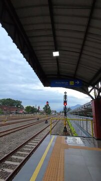 Vertical video of Train station platform under a metal roof. Red signal lights indicate train movement stopped. Yellow safety rails line the platform edge. Blue sign directs passengers to exit. 4K Foo