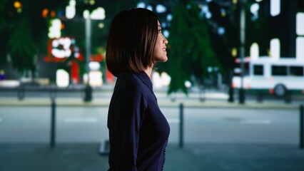 Fototapeta premium Asian person walking around town under streetlights, enjoying urban promenade near city center district. Young woman relaxing on stroll at nighttime, admiring downtown and skyscrapers. Handheld shot.