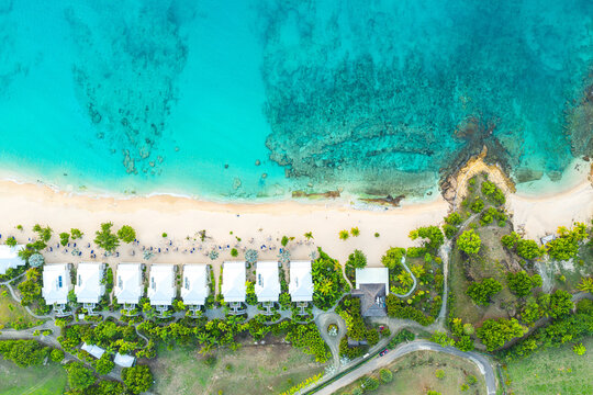Aerial view of luxury beachfront bungalows along a white sandy shore with crystal clear turquoise water and coral reefs English Harbour, Saint Paul, Antigua and Barbuda.