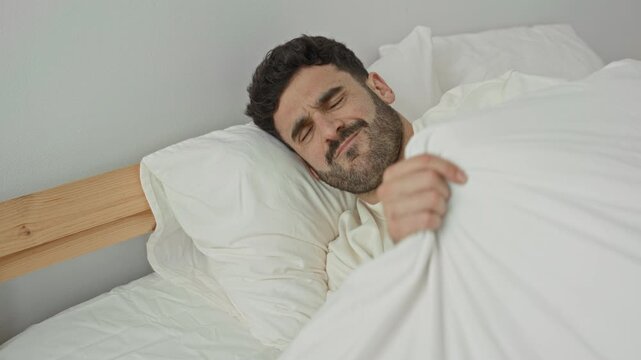 Man pulling duvet over face with hand on bed and resting head on pillow amid white sheets in building; sleepiness rest.