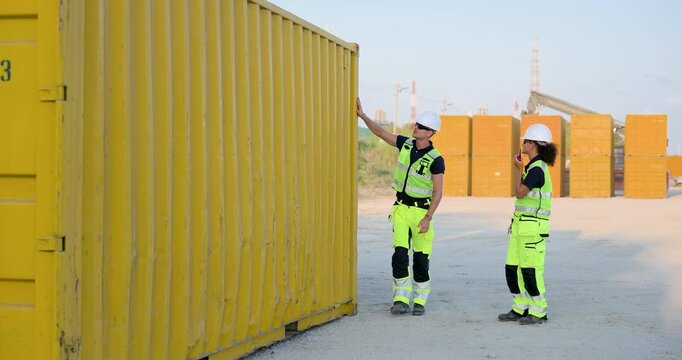 Adult Caucasian woman and man logistics crew inspect yellow container wall, using radio and laptop for cargo verification, yard communication, safety compliance, dispatch planning work zone check