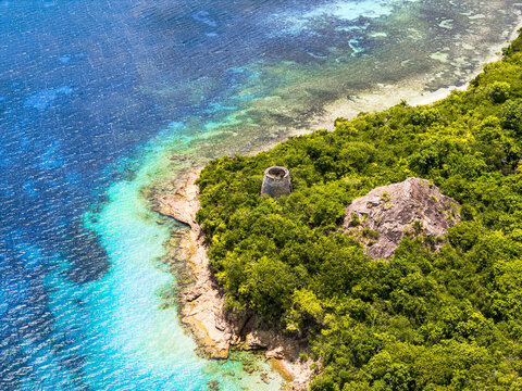Aerial view of a historic stone tower on a lush green coastline with turquoise water and coral reefs in English Harbour, Saint Paul, Antigua and Barbuda.