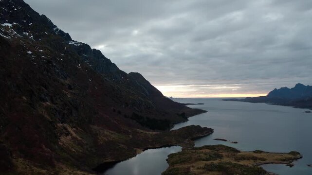 Wide shot of an ocean flowing into a fjord with mountains on either side, under mostly gray clouds with sunlight peering through the distant horizon.