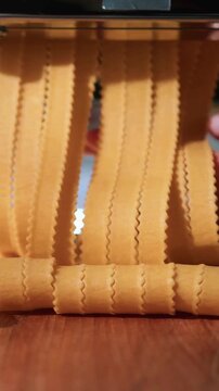 A metal pasta machine cuts sheets of fresh yellow dough into wide ribbons with wavy zig-zag edges.