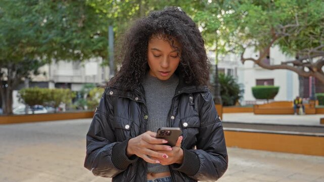 Teenage woman with curly hair holding smartphone, thumbs typing while standing on a city street plaza with trees and benches; urban joy.