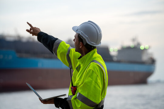 Marine engineer holding a laptop and pointing towards a container ship at a commercial port terminal. Concept of smart port management, digital maritime logistics, and supply chain