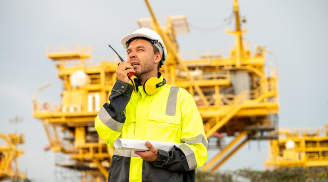 Industrial engineer using a walkie-talkie and holding a rolled blueprint at an offshore energy platform facility. Concept of industrial safety compliance, power infrastructure operations