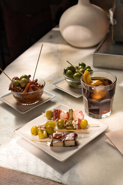 Variety of traditional spanish pintxos and tapas, including seafood skewers, anchovies, cheese, and olives, served on a bar counter with a glass of vermouth