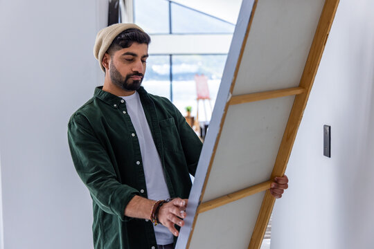 Man wearing knit beanie holding large stretched canvas, revealing wooden stretcher bars in studio