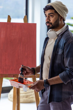 Man standing in studio holding paint tray beside red canvas at easel, wearing beanie, headphones