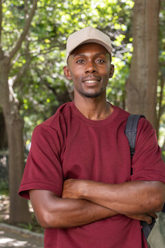 African adult man standing at shaded park, crossing arms, wearing maroon tee, beige cap, backpack