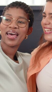 Vertical video: Talking left woman with glasses leading friend in blazer posing for camera on sofa