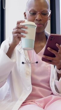Vertical video: Smiling clinician in pink scrubs, steth reading phone on clinic break, sipping cup