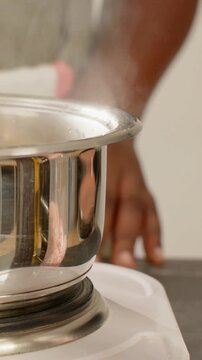Vertical video: Steam rising, man in grey shirt stirring pot on hot plate with ladle preparing food