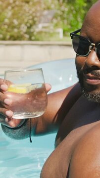 Vertical video: Cam filming shirtless man in shades raising glass, lemon in backyard pool toasting