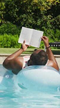 Vertical video: Holding paperback, man floating in tank top reading on ring in pool, turning pages