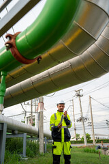 Naklejka premium industrial engineer wearing a white hard hat and high-visibility PPE holds a rolled blueprint and points while speaking on a walkie-talkie by large outdoor refinery pipeline infrastructure.