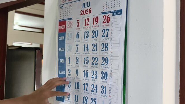 Hands flipping through a May 2026 wall calendar to show the month of June, representing effective time management, planning, and organizing a busy schedule in a home office or business environment.