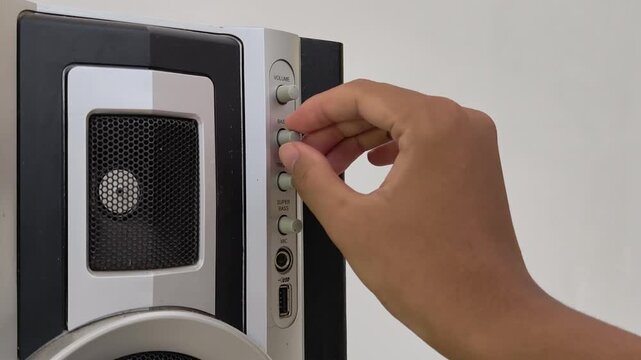 Close-up of a hand turning a dial on an electronic stereo speaker. The silver control panel features knobs for volume and bass, alongside audio inputs. The image depicts manual adjustment of sound set