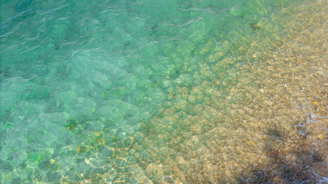 Crystal clear turquoise sea water ripple surface with colorful pebble stones under the shallow beach wave. Natural background texture of clean tropical ocean shore during summer holiday vacation.