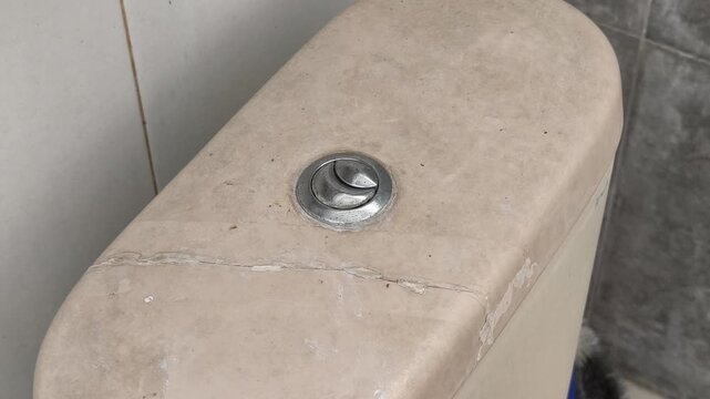 Close up of a dirty and cracked beige ceramic toilet tank with a dual flush chrome button in an old bathroom, showing signs of neglect and wear needing repair or replacement.