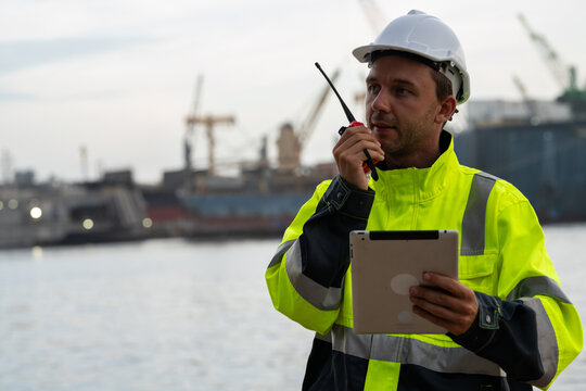 Marine engineer using a digital tablet to inspect fleet operations at a commercial port terminal with a blurred cargo ship background. Concept of smart logistics and supply chain technology.