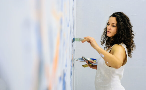 Female artist painting a large wall mural in a bright studio while holding brushes and palette. Creative process, concentration and contemporary visual art in progress