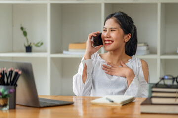 Happy young Asian businesswoman talking on smartphone while working with laptop at office desk. © Songsak C
