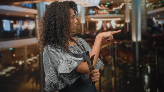 Woman waitress with curly hair wearing apron, holding cutlery and pointing finger toward a table in a building dining area; service determination.