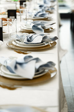 Beautifully styled table setting for a formal event with a white tablecloth, gold charger plates, white dinnerware, blue gradient napkins, clear water glasses and golden cutlery.