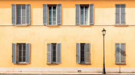 Obraz premium European building facade presenting a vibrant yellow wall with multiple windows, open and closed grey shutters, and a vintage street lamp