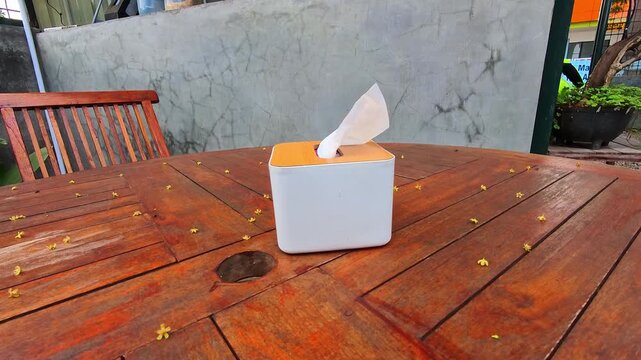 Video of tissue paper blowing in the wind inside a tissue box sitting on a warm wooden table. The paper napkin slightly protrudes from the top. Tiny yellow petals scatter around the tabletop. 4K Foota