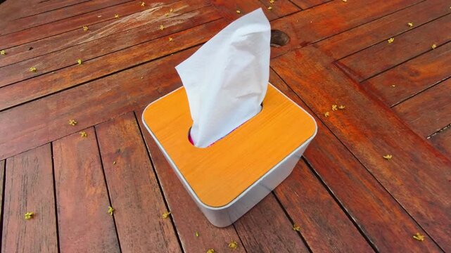 Video of tissue paper blowing in the wind inside a tissue box sitting on a warm wooden table. The paper napkin slightly protrudes from the top. Tiny yellow petals scatter around the tabletop. 4K Foota