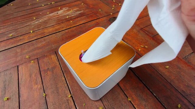Vertical Footage of a hand picking up a tissue in a box. Tissue box with orange top and white edges. Tissue pulled out, slightly crumpled, mid-use. Modern, clean design sits on warm-toned planks. 4K V