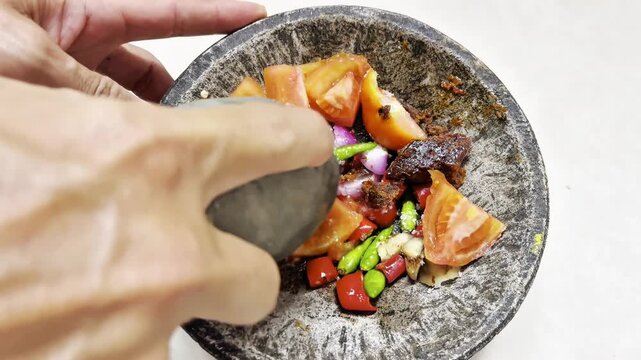 Close-up footage of traditional Indonesian sambal being prepared with fresh tomatoes, chili peppers, onions, and spices using a stone mortar and pestle. Authentic culinary process showcasing homemade 