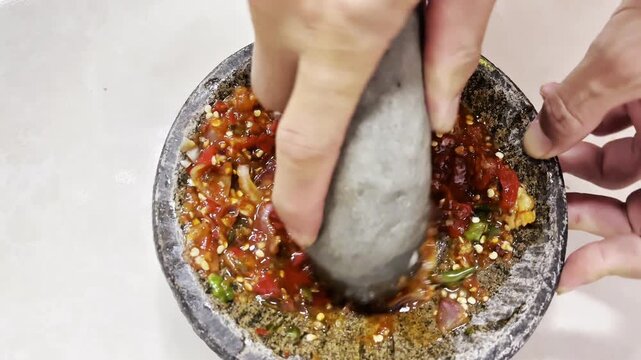 Hands grinding fresh ingredients in a stone mortar to make Indonesian chili sauce. Authentic food preparation video featuring tomatoes, chilies, shallots, and traditional kitchen methods.